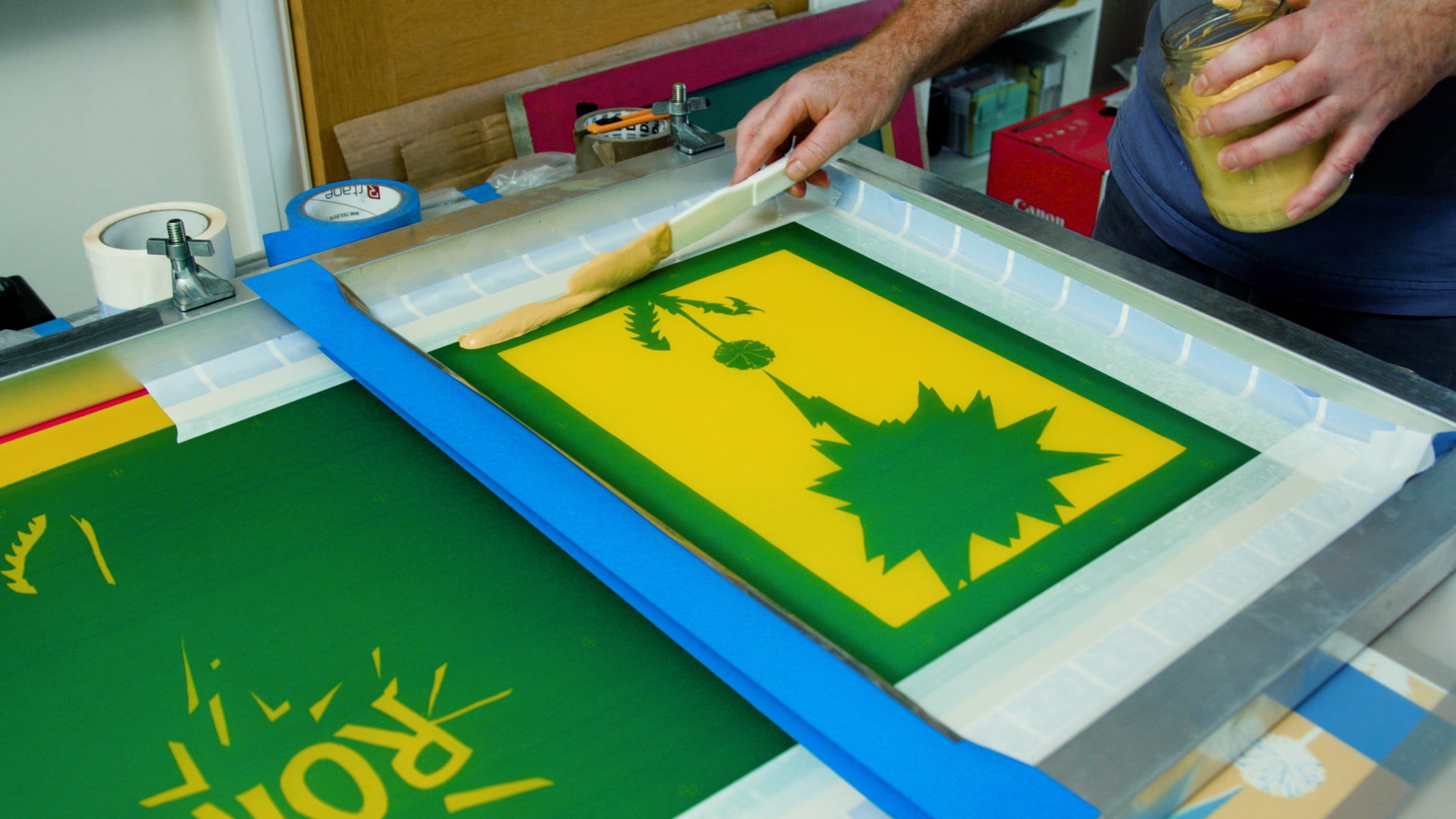 Applying the colour background paint to the screen, before running the squeegee over the silkscreen. The screen is set up with tape to stop the paint moving around the screen